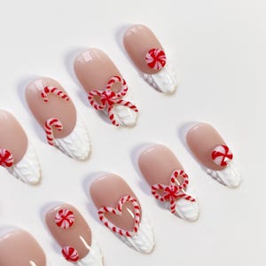 May include: Oval-shaped artificial nails with a light pink base and white tips. Each nail features a festive design, including red and white candy canes, bows, and heart shapes. The nails have a textured, white base.