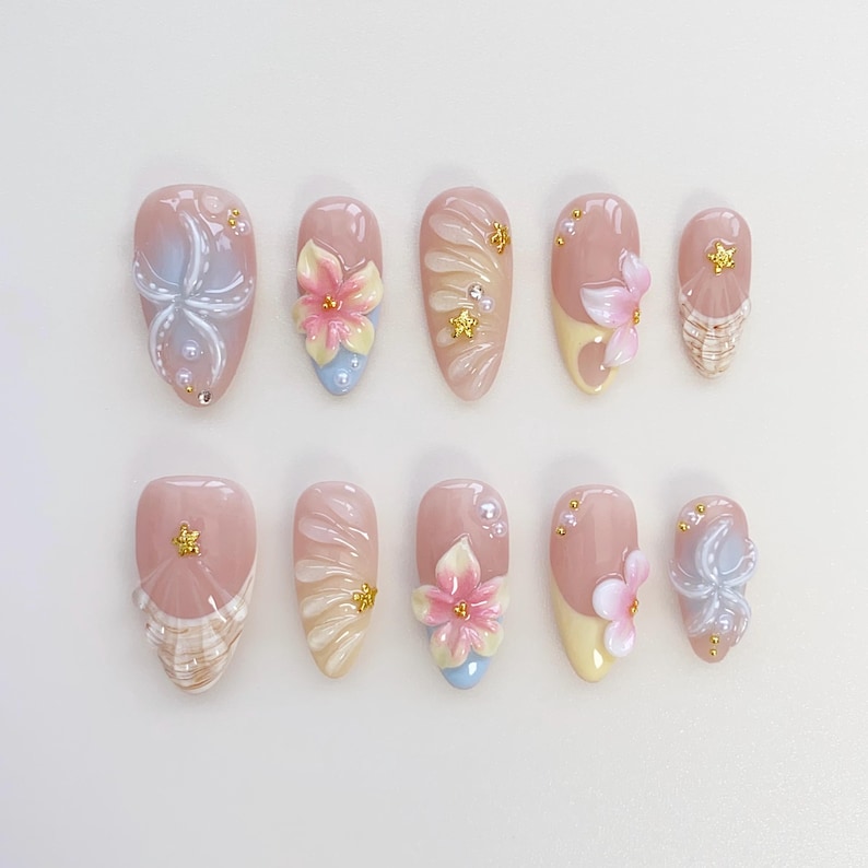 May include: A set of ten artificial nails in various designs. The nails feature a light pink base with floral and abstract designs in white, blue, and pink. Some nails have 3D flower accents and gold embellishments. The nails are oval-shaped.