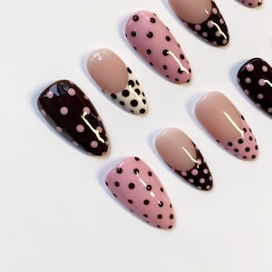 Peut inclure: Collection de faux ongles aux designs vari&eacute;s. Certains sont rose clair &agrave; pois noirs, d'autres marron fonc&eacute; &agrave; pois roses, et certains ont une base nude avec un motif &agrave; pois noir et blanc.
