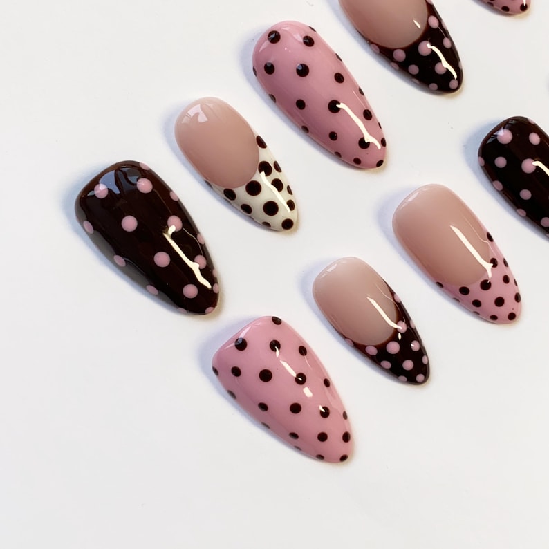 Peut inclure: Collection de faux ongles aux designs vari&eacute;s. Certains sont rose clair &agrave; pois noirs, d'autres marron fonc&eacute; &agrave; pois roses, et certains ont une base nude avec un motif &agrave; pois noir et blanc.