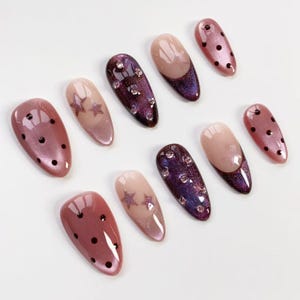 May include: A collection of ten artificial nails in various designs. Some nails are a mauve color with black polka dots, others feature star designs, and some have a dark purple base with rhinestones.