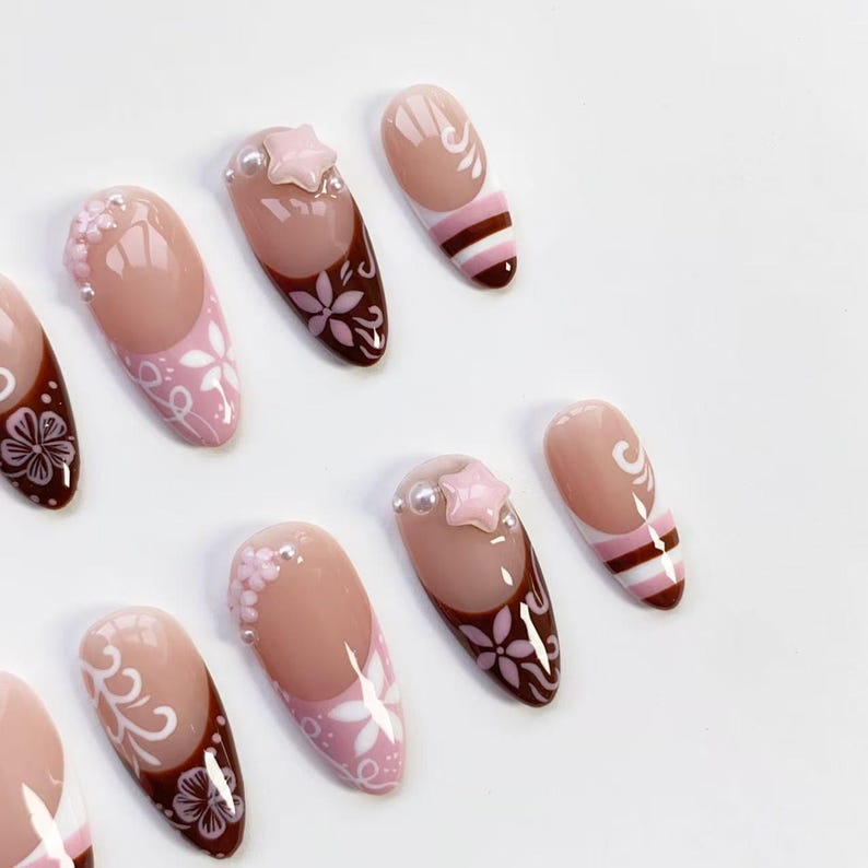 May include: A collection of oval-shaped false nails. The nails feature a variety of designs, including floral patterns, star-shaped embellishments, and striped accents. The color palette includes shades of pink, brown, and white, creating a delicate and feminine aesthetic.