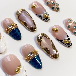 May include: A collection of artificial nails in various designs. Some nails feature a nude base with gold accents, while others have a blue textured tip. Several nails have gold embellishments and some have a zebra print design.