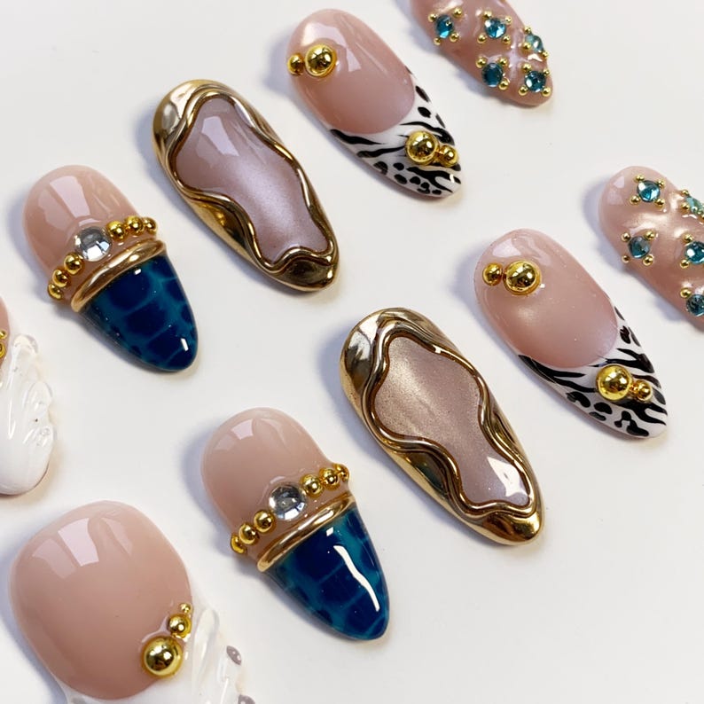 May include: A collection of artificial nails in various designs. Some nails feature a nude base with gold accents, while others have a blue textured tip. Several nails have gold embellishments and some have a zebra print design.