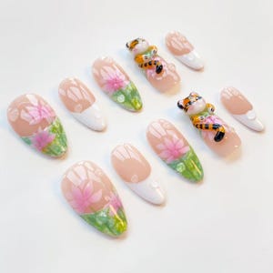 May include: A collection of artificial nails with various designs. Some nails feature a pink floral design with green accents, while others have a tiger figurine. Several nails have a white tip.