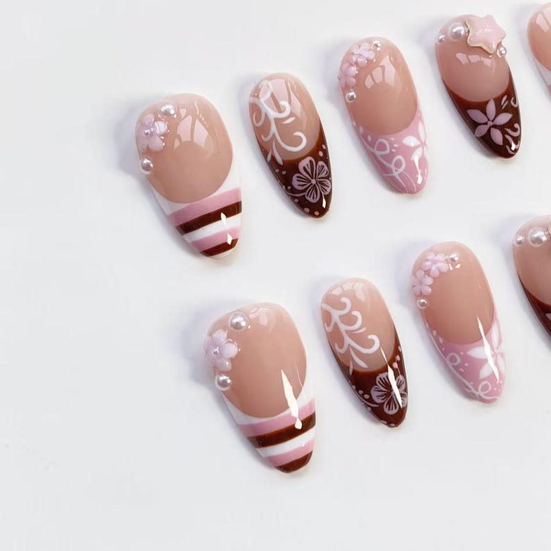 May include: A collection of artificial nails with various designs. Some nails feature a nude base with white and brown floral patterns, while others have a nude base with white and brown stripes. Some nails have pearl accents.