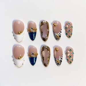 May include: A set of ten artificial nails in various designs. Some nails feature a nude base with gold accents, while others have a white tip with gold details. Other nails have a gold frame with a nude center, and some have blue gems.