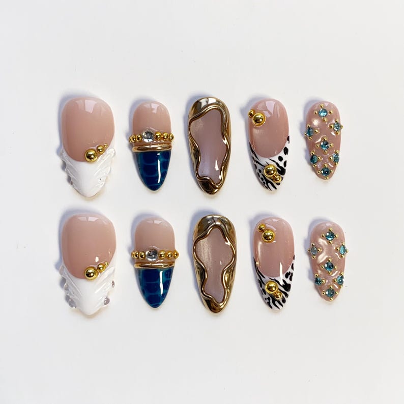 May include: A set of ten artificial nails in various designs. Some nails feature a nude base with gold accents, while others have a white tip with gold details. Other nails have a gold frame with a nude center, and some have blue gems.