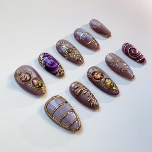 Pode incluir: Um conjunto de dez unhas artificiais cor de lavanda com enfeites dourados. Os designs incluem luas, s&oacute;is, estrelas e padr&otilde;es abstratos. Algumas unhas apresentam pedras preciosas iridescentes. As unhas est&atilde;o dispostas sobre uma superf&iacute;cie branca.