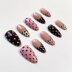 Peut inclure: Un ensemble de dix faux ongles de diff&eacute;rentes conceptions. Certains ongles sont roses unis avec des pois noirs, tandis que d'autres ont une base rose avec des pois noirs ou une couleur marron fonc&eacute; unie avec des pois roses. Certains ongles ont une pointe nude.