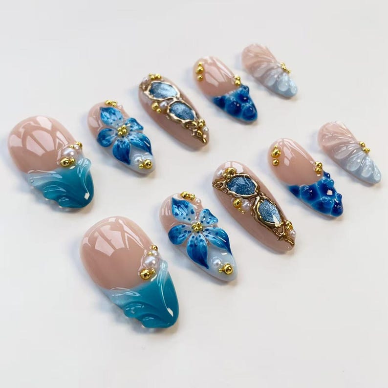 May include: A collection of artificial nails with various designs. Some nails feature a nude base with blue floral accents, while others showcase a blue and white wave design. Several nails have gold and pearl embellishments.