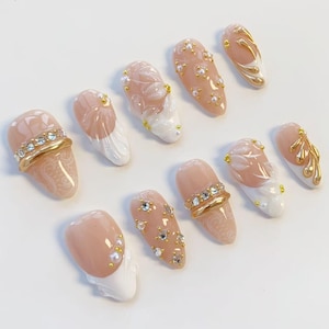 May include: A collection of ten artificial nails in various designs. The nails feature a nude base color with white, gold, and clear accents. Some nails have pearl and rhinestone embellishments, while others showcase intricate floral patterns.