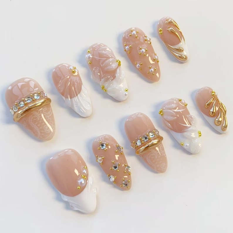 May include: A collection of ten artificial nails in various designs. The nails feature a nude base color with white, gold, and clear accents. Some nails have pearl and rhinestone embellishments, while others showcase intricate floral patterns.
