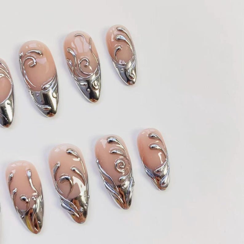 May include: A set of almond-shaped artificial nails. Each nail features a pale pink base with a silver metallic design. The silver design includes a swirl pattern and a metallic tip, creating a glamorous look.