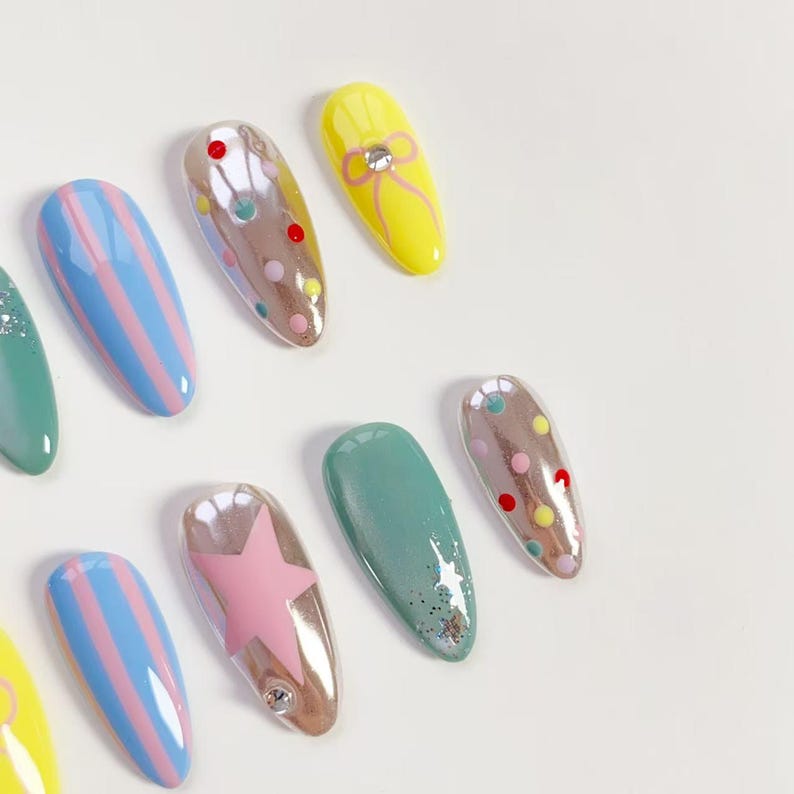 K&ouml;nnte beinhalten: Eine Sammlung farbenfroher, mandelf&ouml;rmiger Kunstn&auml;gel. Designs umfassen pastellblaue und rosa Streifen, einen rosa Stern, Gelb mit einer Schleife und schillernde N&auml;gel mit Tupfen. Einige N&auml;gel haben Glitzerakzente. Die N&auml;gel sind auf einer wei&szlig;en Oberfl&auml;che angeordnet.