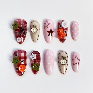 Puede incluir: Un conjunto de diez uñas artificiales con varios diseños. Algunas uñas presentan un patrón de cuadros rojos, mientras que otras son de color rosa o beige liso. Las decoraciones incluyen estrellas, botones y pequeños ositos de peluche. Las uñas tienen forma ovalada.