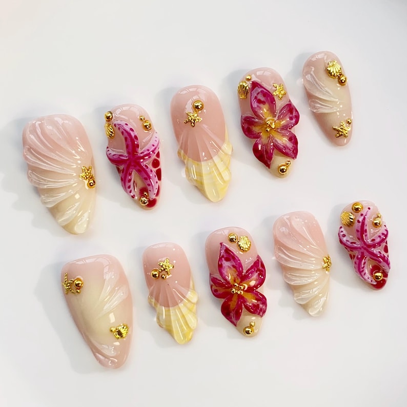 May include: A set of ten artificial nails with a light pink base, featuring various designs. Some nails have a starfish, others have a flower, and some have a seashell design. All nails have gold embellishments.