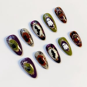 May include: A collection of Halloween-themed false nails. Designs include a spider web with a spider, a bat, a ghost, and a pumpkin. The nails feature a variety of colors, including purple, green, orange, and black, with gold accents.