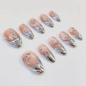 May include: A set of ten almond-shaped false nails. Each nail features a pale pink base with a silver, ornate design and a silver metallic tip. The nails are arranged on a white surface.
