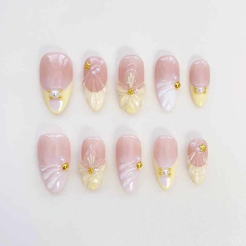 May include: A collection of ten artificial nails with diverse designs. The nails have a light pink base with yellow tips. Some nails feature 3D floral and pearl accents, while others have white and gold embellishments. The nails are oval-shaped.