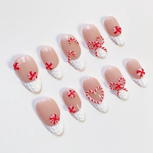 May include: Set of ten artificial nails with a light pink base and white textured tips. Each nail features red and white candy cane or bow designs, perfect for festive holiday wear.