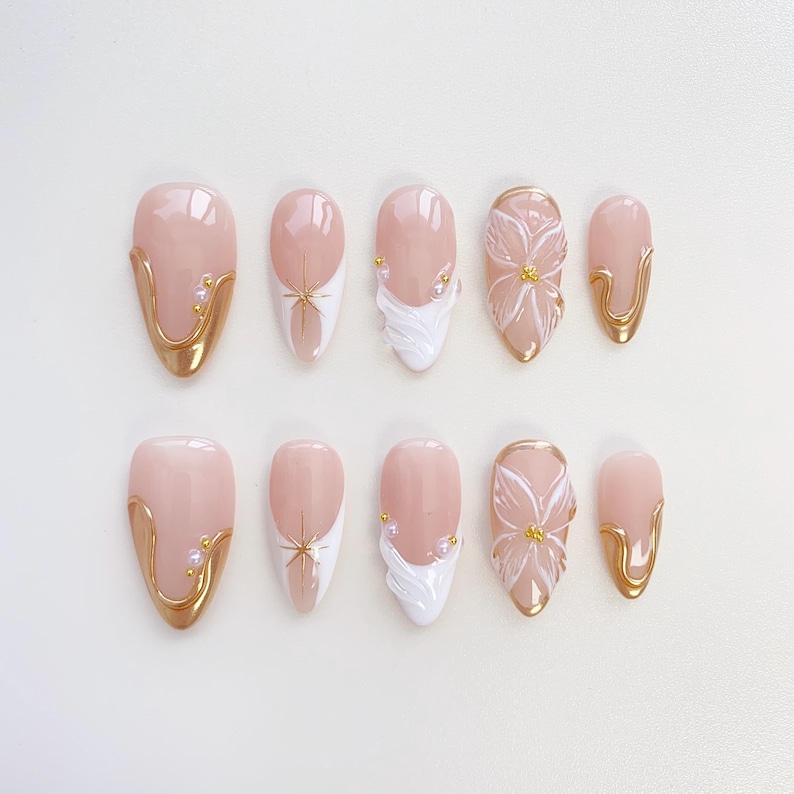May include: A set of ten almond-shaped false nails. The nails feature a pale pink base with gold accents, white designs, and floral embellishments. Some nails have a gold border, while others have a starburst or a flower design.