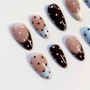Puede incluir: Una colecci&oacute;n de u&ntilde;as postizas de forma ovalada. Las u&ntilde;as presentan una variedad de dise&ntilde;os, incluyendo marr&oacute;n oscuro liso, rosa claro con lunares marrones oscuros y rosa claro con punta azul claro y lunares marrones oscuros. Las u&ntilde;as est&aacute;n dispuestas sobre una superficie blanca.