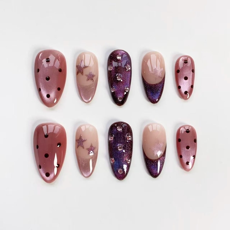 May include: A set of ten artificial nails in various designs. The nails feature a mix of colors, including pink, purple, and nude. Some nails have black polka dots, star accents, or sparkling rhinestones. The nails are oval-shaped.