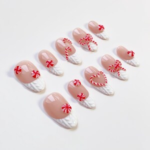 May include: A set of ten artificial nails with a light pink base and white textured tips. Each nail is decorated with red and white candy cane or peppermint designs, including bows, hearts, and circular shapes.