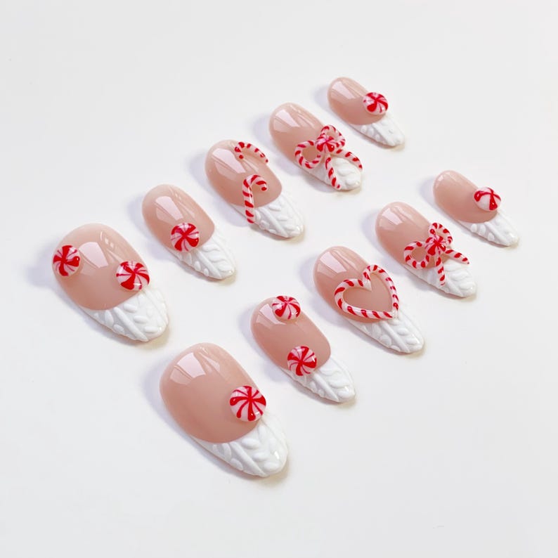 May include: A set of ten artificial nails with a light pink base and white textured tips. Each nail is decorated with red and white candy cane or peppermint designs, including bows, hearts, and circular shapes.