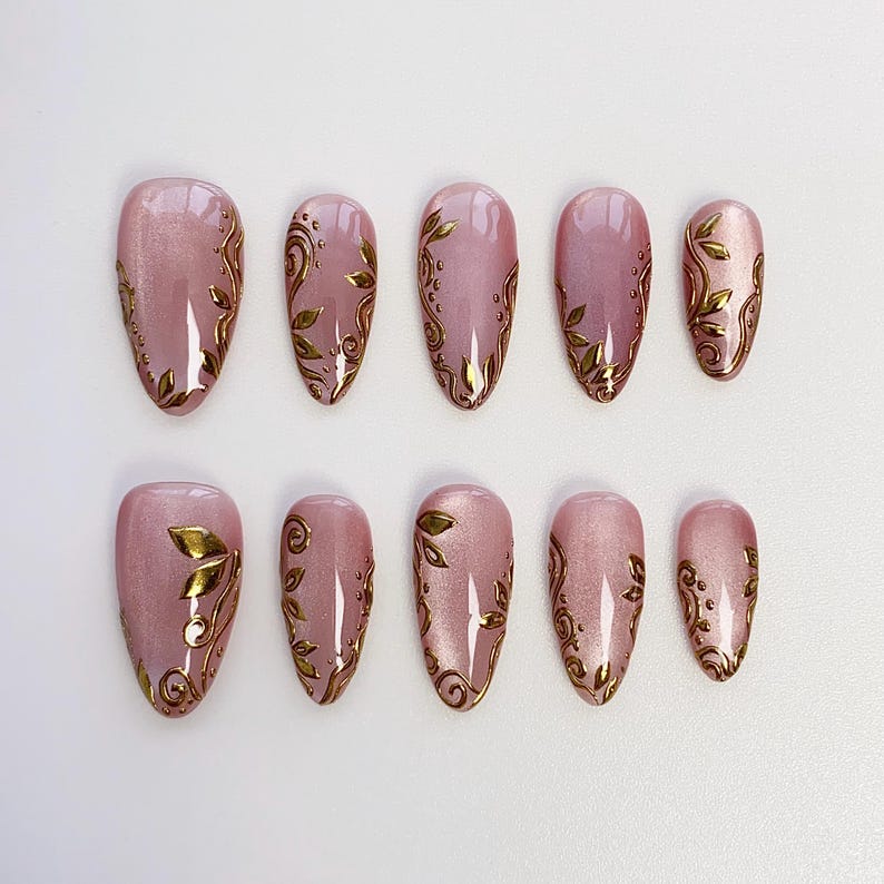 May include: Set of ten almond-shaped false nails. Each nail is a pale pink with a gold leaf design. The nails are arranged in two rows of five, showcasing the varying sizes.