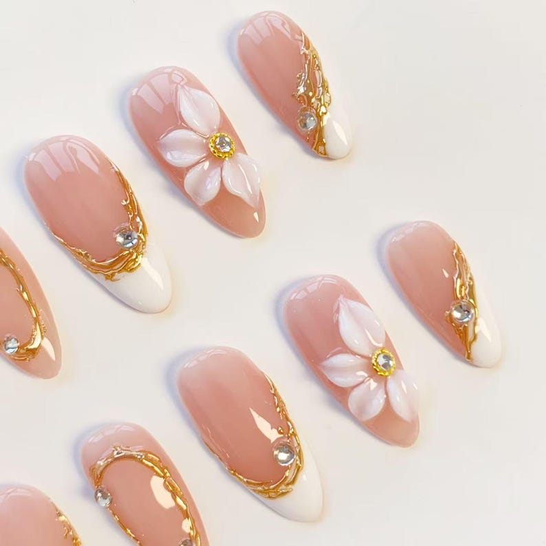 Puede incluir: Un juego de u&ntilde;as postizas en forma de almendra con base nude y puntas blancas. El dise&ntilde;o incluye remolinos met&aacute;licos dorados en relieve, pedrer&iacute;a transparente y p&eacute;talos de flores blancos en 3D con centros dorados en algunas u&ntilde;as.
