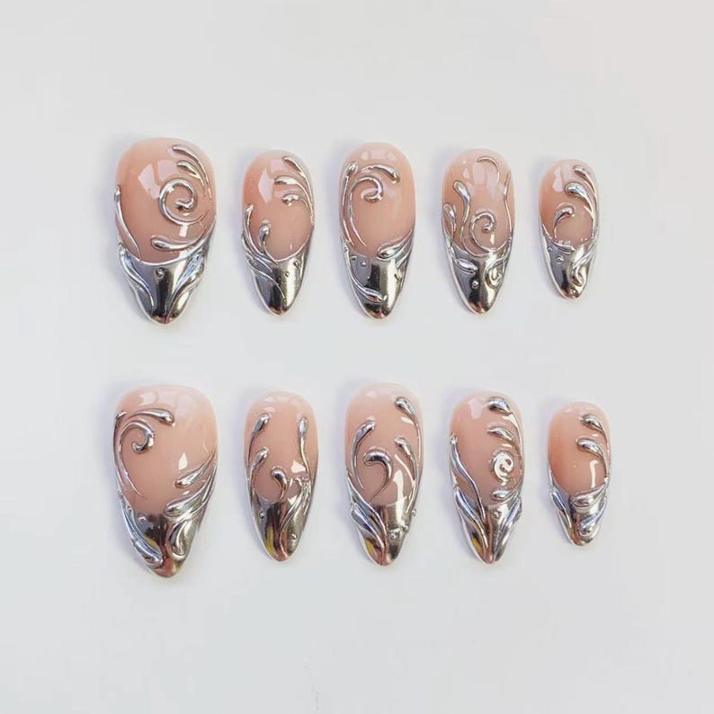 May include: Set of ten artificial nails in various sizes. Each nail features a nude-colored base with a silver metallic design. The silver design includes a swirl pattern and a metallic tip, creating a decorative and elegant look.