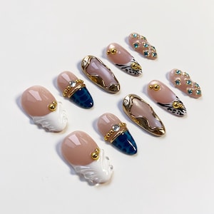 May include: A collection of artificial nails in various designs. Some nails feature a nude base with gold accents, while others have a blue textured pattern. Several nails have a gold border with a pinkish-beige center.