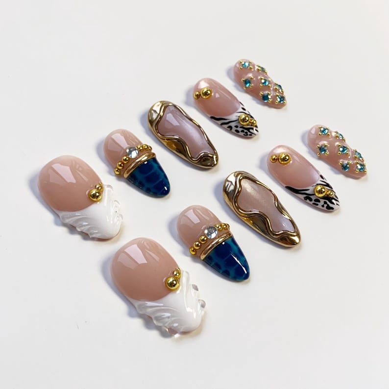 May include: A collection of artificial nails in various designs. Some nails feature a nude base with gold accents, while others have a blue textured pattern. Several nails have a gold border with a pinkish-beige center.