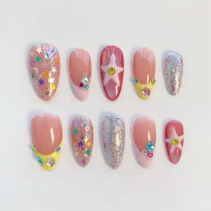 Pu&ograve; includere: Un set di dieci unghie finte con vari design. Le unghie presentano una gamma di colori, tra cui rosa, argento e rosso, con abbellimenti come glitter, stelle e gemme. Alcune unghie hanno un design a punta francese con accenti gialli o rosa.
