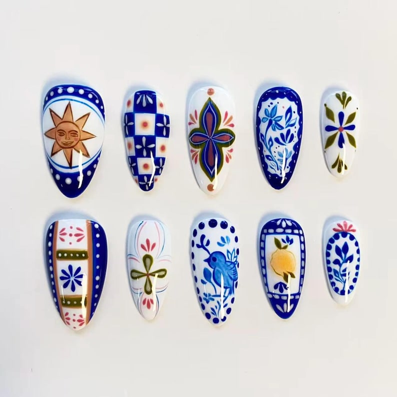 May include: A set of ten almond-shaped press-on nails featuring a variety of hand-painted designs in blue, white, yellow, and green. Patterns include a sun face, checkered squares, floral motifs, a small blue bird, and a lemon illustration.