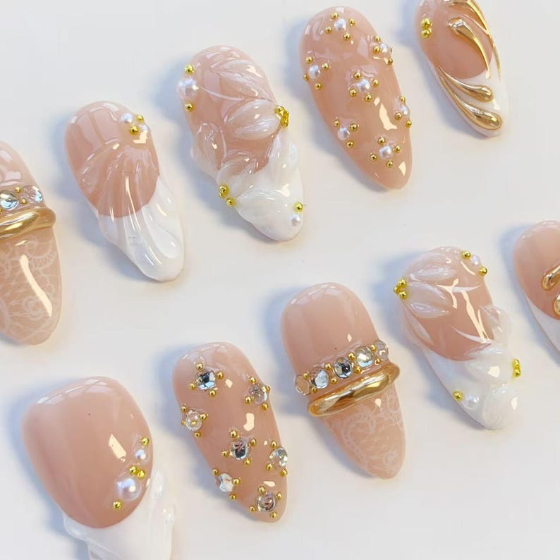 May include: A collection of artificial nails in various designs. Some nails are nude-colored with white floral accents, gold embellishments, and small pearls. Others feature gold rings with rhinestones or lace patterns.