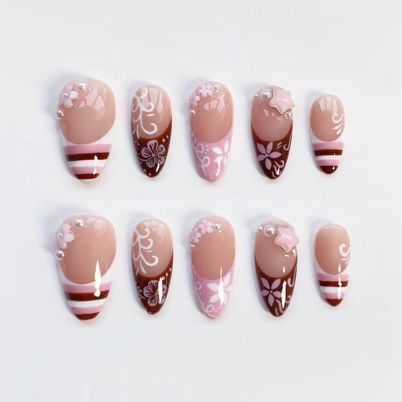 May include: A set of ten artificial nails in various designs. The nails feature a nude base with accents of pink, white, and brown. Some nails have floral patterns, pearls, or star-shaped embellishments. Others have striped or swirl designs.