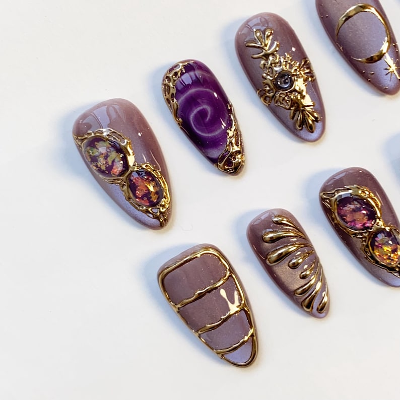 Pode incluir: Close-up de um conjunto de unhas artificiais com v&aacute;rios designs. As unhas apresentam uma cor de base malva com enfeites dourados, incluindo redemoinhos, padr&otilde;es florais e detalhes de pedras preciosas. As unhas est&atilde;o dispostas sobre uma superf&iacute;cie branca.