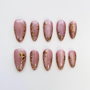 May include: A set of ten almond-shaped, press-on nails. Each nail is a soft pink with a gold leaf design. The gold leaf design varies on each nail, with some featuring full leaf patterns and others with delicate swirls.