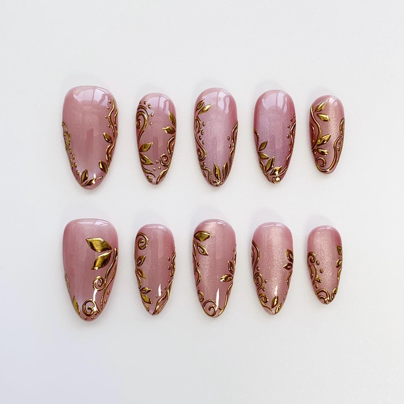 May include: A set of ten almond-shaped, press-on nails. Each nail is a soft pink with a gold leaf design. The gold leaf design varies on each nail, with some featuring full leaf patterns and others with delicate swirls.