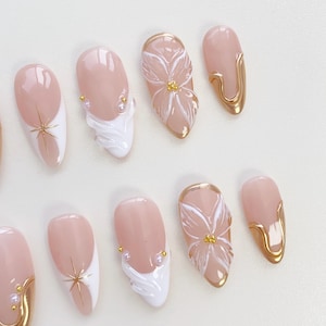 May include: A selection of artificial nails with diverse designs. Some nails have a nude base with white tips and gold star details. Others display white floral patterns, gold trim, and pearl embellishments. The nails are oval-shaped.