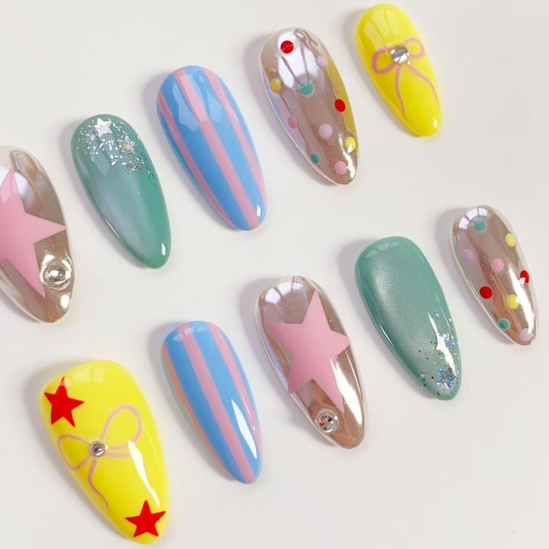 K&ouml;nnte beinhalten: Eine Sammlung k&uuml;nstlicher N&auml;gel mit verschiedenen Designs. Einige N&auml;gel sind gelb mit roten Sternen und einer Schleife, andere blau mit rosa Streifen, und einige haben rosa Sterne oder bunte Punkte. Die N&auml;gel sind oval und haben ein gl&auml;nzendes Finish.