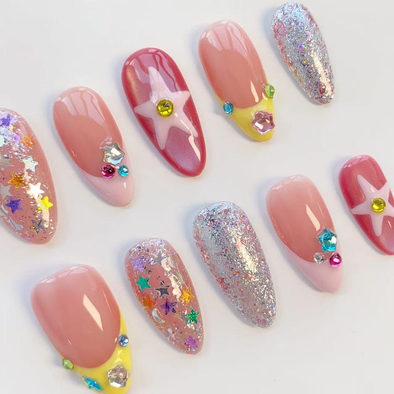 Pu&ograve; includere: Una collezione di unghie finte con vari design. Alcune unghie presentano glitter, decorazioni a stella e gioielli. Altre hanno un design a punta francese rosa e gialla. Le unghie ovali sono disposte su uno sfondo bianco.