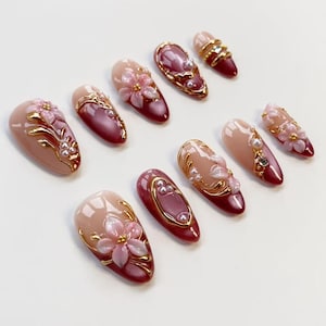 May include: A collection of artificial nails in various designs. The nails feature a gradient of nude and burgundy colors, adorned with gold accents, pearls, and 3D floral embellishments. Some nails have a gold border, while others have a pearl accent.