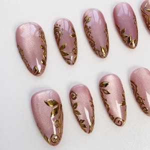 May include: A set of oval-shaped, light pink artificial nails with gold leaf and swirl designs. The nails are arranged on a white surface, showcasing the intricate details of the nail art. The gold accents provide a contrast to the pink base.