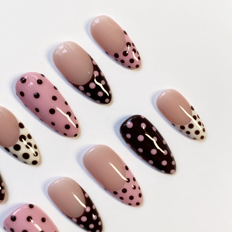 Peut inclure: Une collection de faux ongles en forme d'amande. Certains sont rose clair avec des pois noirs, d'autres sont marron fonc&eacute; avec des pois roses. Plusieurs ongles ont une base nude avec un motif &agrave; pois marron fonc&eacute;.
