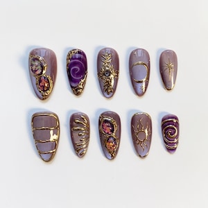 Pode incluir: Um conjunto de dez unhas artificiais com v&aacute;rios designs. As unhas s&atilde;o principalmente lavanda com detalhes dourados e enfeites. Algumas unhas apresentam pedras preciosas, enquanto outras exibem motivos celestes como luas, estrelas e s&oacute;is. A est&eacute;tica geral &eacute; ornamentada e elegante.