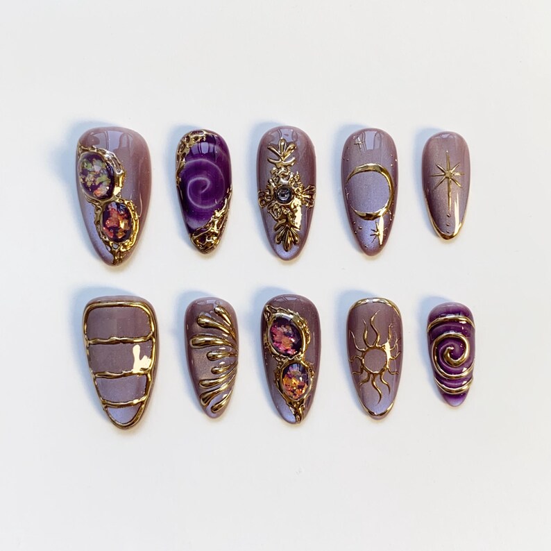 Pode incluir: Um conjunto de dez unhas artificiais com v&aacute;rios designs. As unhas s&atilde;o principalmente lavanda com detalhes dourados e enfeites. Algumas unhas apresentam pedras preciosas, enquanto outras exibem motivos celestes como luas, estrelas e s&oacute;is. A est&eacute;tica geral &eacute; ornamentada e elegante.
